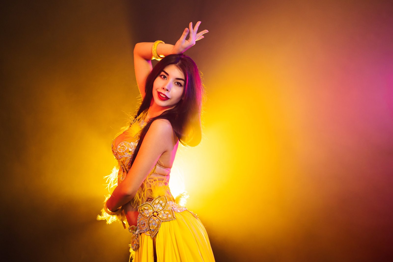Seductive sexy traditional oriental belly dancer girl dancing on yellow neon smoke background. Woman in exotic costume with feathers sexually moves her semi-nude body.